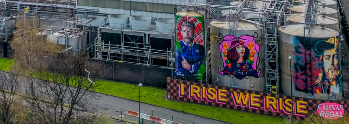 Aerial view of Strathclyde grain distillery in Glasgow's Gorbals, showing its production buildings, cooling towers, storage tanks and large painted murals including the 'I Rise We Rise' artwork and Chivas Regal branding.