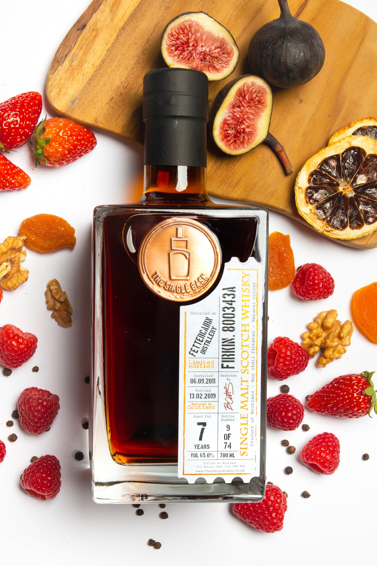 A bottle of Fettercairn whisky with a wooden cap and label, surrounded by assorted fruits and nuts on a white surface.